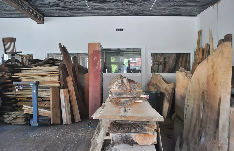 Holzlager einer Werkstatt mit gestapelten Brettern, aufrecht stehenden Bohlen und Baumscheiben mit Naturkante, hinten ein Fenster zur Werkstatt.