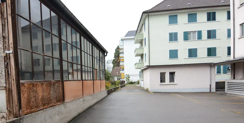 Seitenwand einer ehemaligen Hinterhoftischlerei mit verglaster Fensterfront in Holzrahmen, dahinter ein Hof mit Wohnhäusern in Zürich-Altstetten.