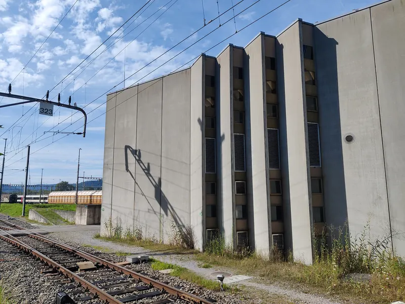 Werkhalle aus grauem Beton mit senkrechten Fensterbändern neben einem Gleis, darüber Fahrleitungen und blauer Himmel mit Wolken.